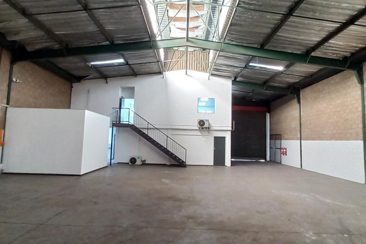 Spacious industrial warehouse featuring high ceilings, exposed steel trusses, a mezzanine level with staircase, roller shutter door, and concrete flooring.