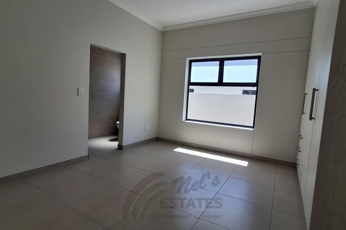 A bright, modern bedroom featuring neutral floor tiles, built-in cupboards, and an en-suite bathroom entrance.