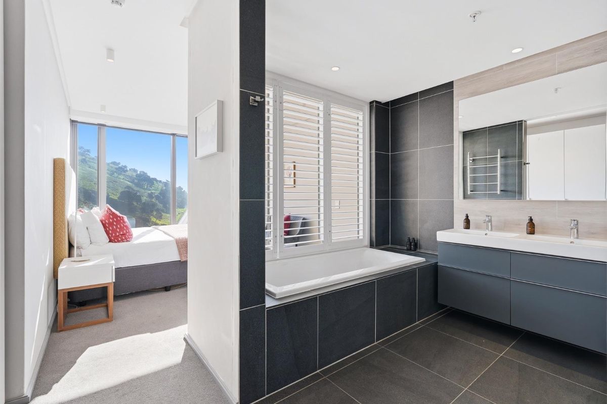 Modern open-plan bathroom featuring a built-in bathtub, double vanity with sleek cabinetry, and plantation shutters, seamlessly connected to the adjacent bedroom area.