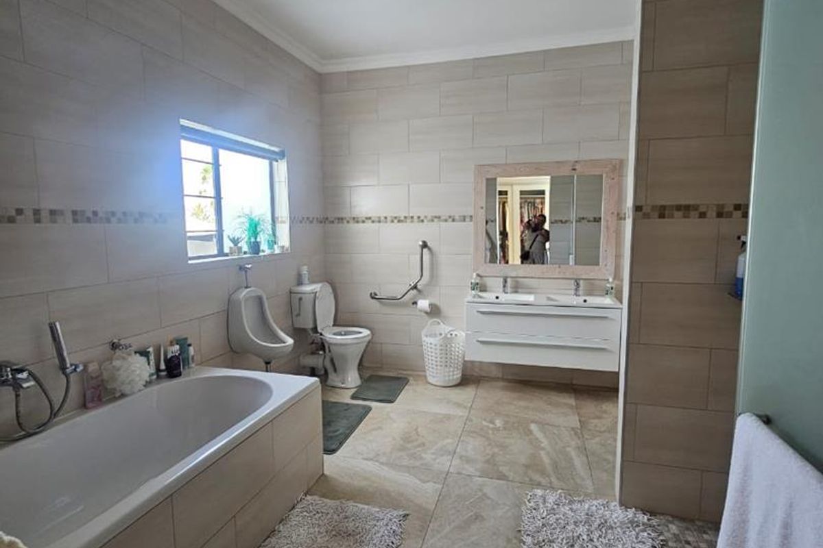 Spacious bathroom featuring a built-in bathtub, wall-mounted vanity with double basins, toilet, and a separate urinal, finished with neutral floor-to-ceiling tiling.