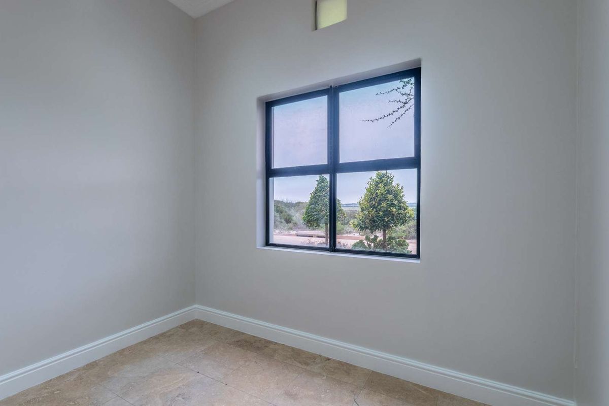 A bright, empty bedroom featuring neutral walls, tiled flooring, and a large black-framed window with a view of the surrounding landscape.
