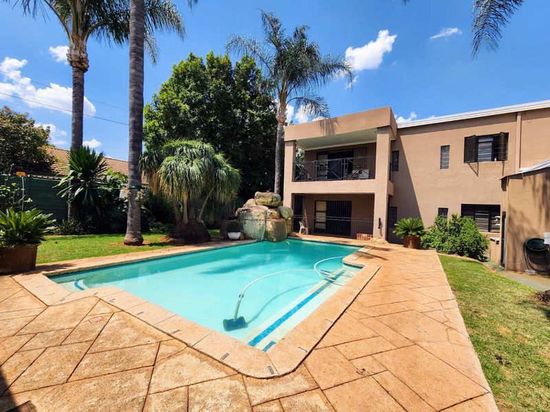 A spacious backyard featuring a swimming pool with a rock water feature, paved surround, and a two-story home with a balcony.