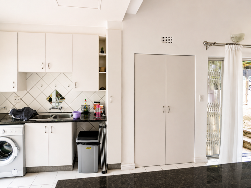 A functional kitchen area featuring white cabinetry, granite countertops, a double basin sink, and an integrated washing machine with direct access to the patio.