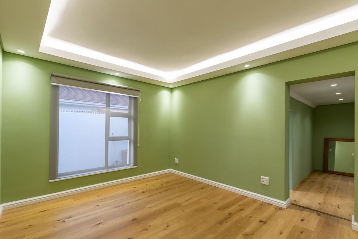 A bright, empty bedroom featuring modern recessed tray ceiling lighting, light wood flooring, and a large window with a roller blind.