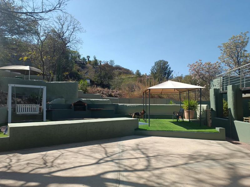 Spacious outdoor entertainment area featuring a built-in braai, tiered retaining walls, concrete paving, and an artificial grass section under a gazebo.