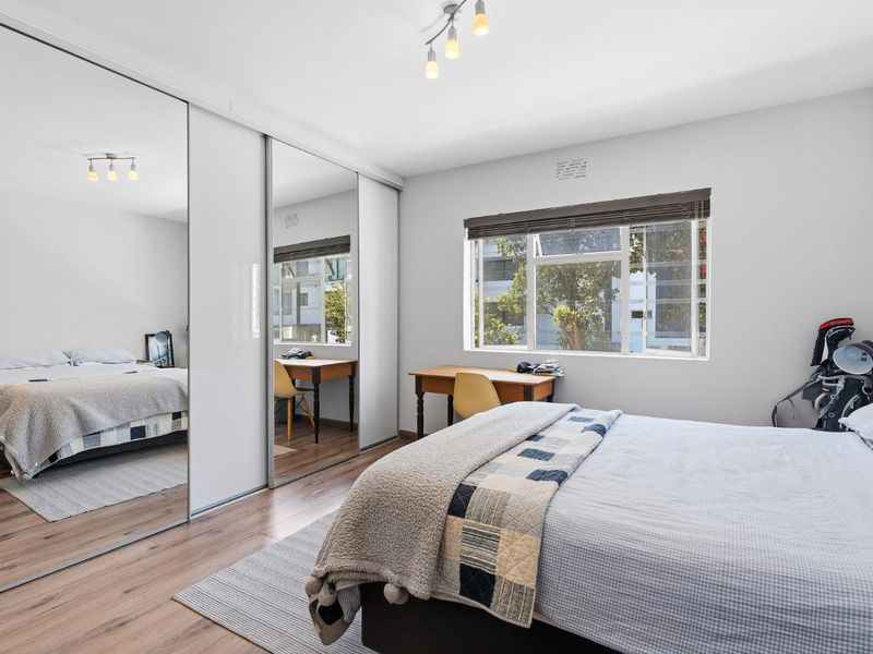 A well-lit bedroom featuring laminate flooring, a large mirrored sliding wardrobe, and a window with a roller blind.