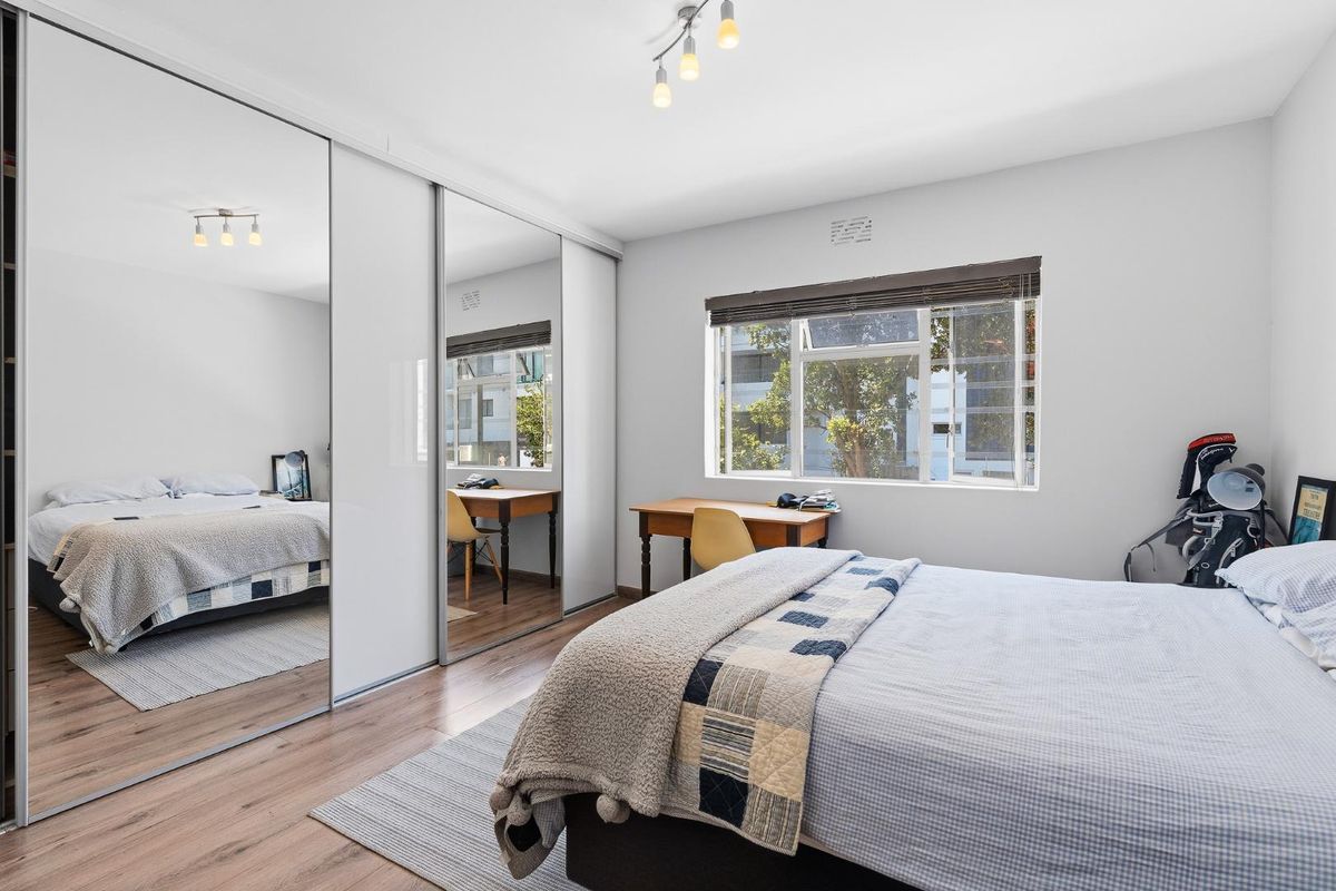 A well-lit bedroom featuring laminate flooring, a large mirrored sliding wardrobe, and a window with a roller blind.