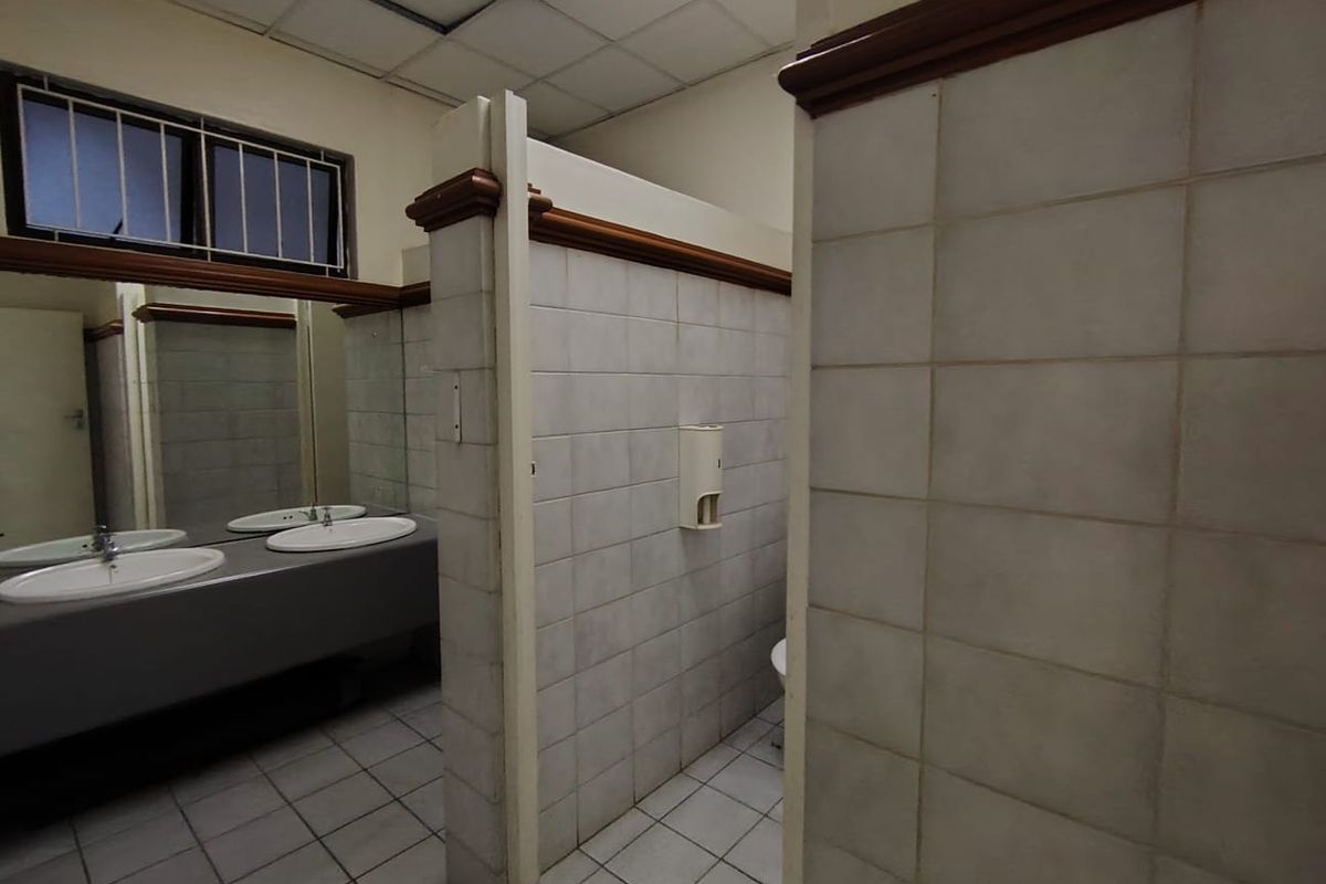 A functional commercial bathroom featuring tiled partitions, built-in vanity with dual sinks, and suspended ceiling tiles, requiring cosmetic updates to modernize the space.