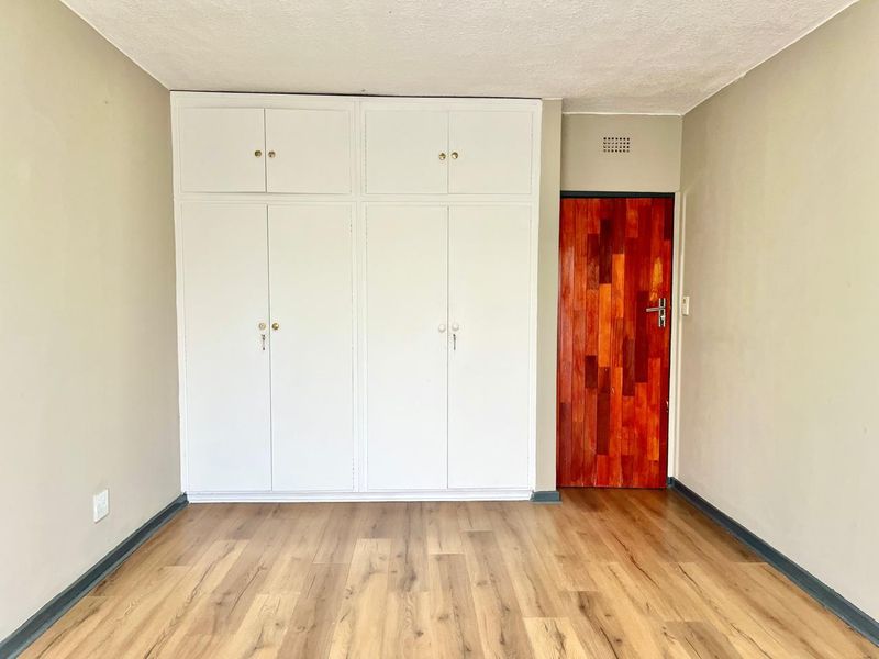 A bright, empty bedroom featuring light wood-look laminate flooring, ample white built-in storage cupboards, and a distinctive wooden door.