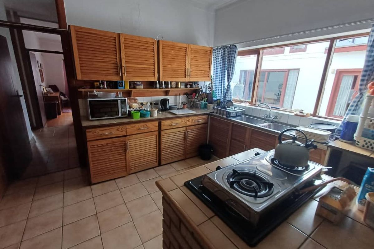 A functional kitchen featuring wooden louvered cabinetry, tiled countertops, a built-in hob, and a double sink with large windows overlooking the exterior.