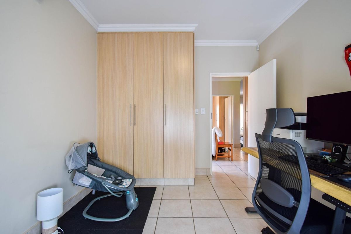 A functional bedroom or office space featuring built-in light wood cabinetry, tiled flooring, and crown molding, with a doorway leading to an adjacent hallway.