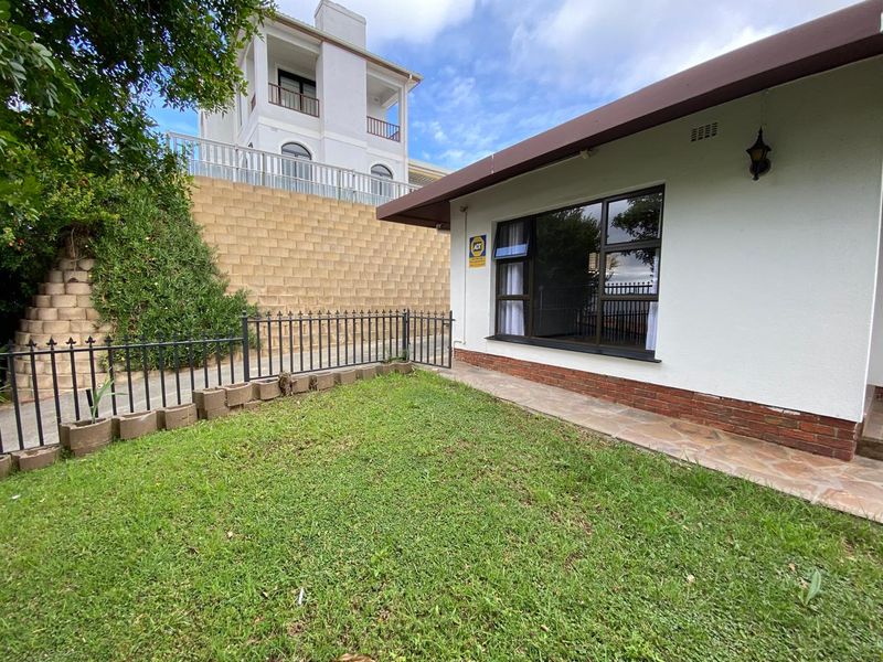 Rear exterior view of the apartment featuring a small lawn, paved walkway, metal fencing, and a retaining wall.