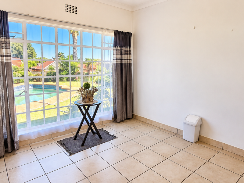 A bright, empty bedroom featuring tiled flooring and a large window with views overlooking the garden and swimming pool area.