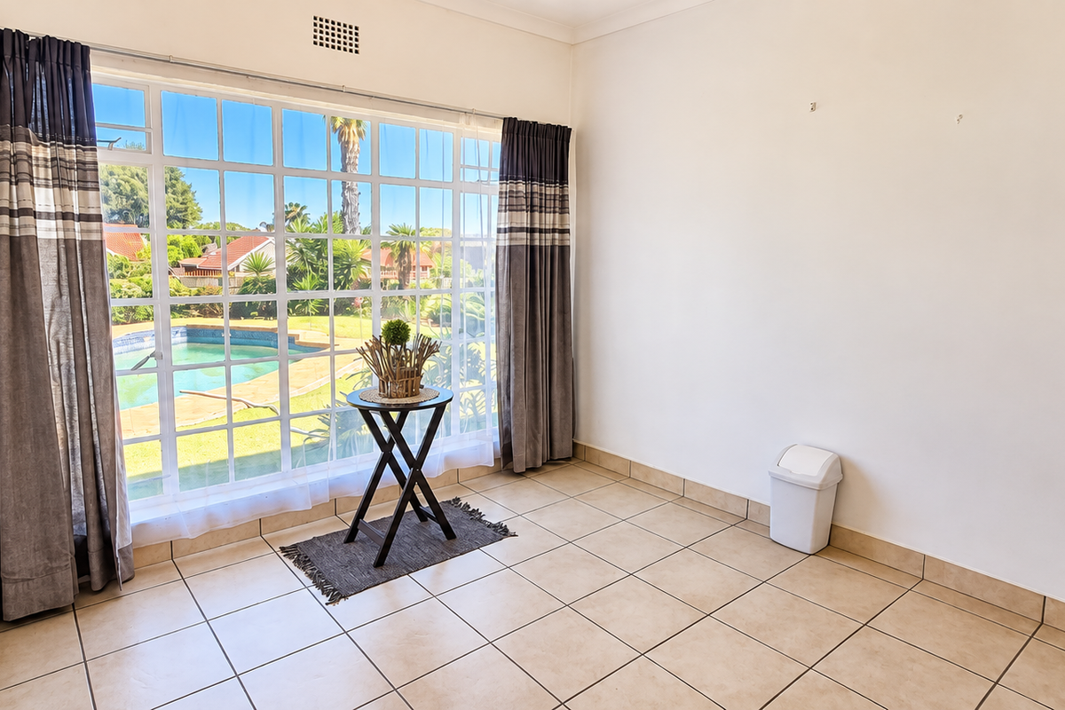 A bright, empty bedroom featuring tiled flooring and a large window with views overlooking the garden and swimming pool area.