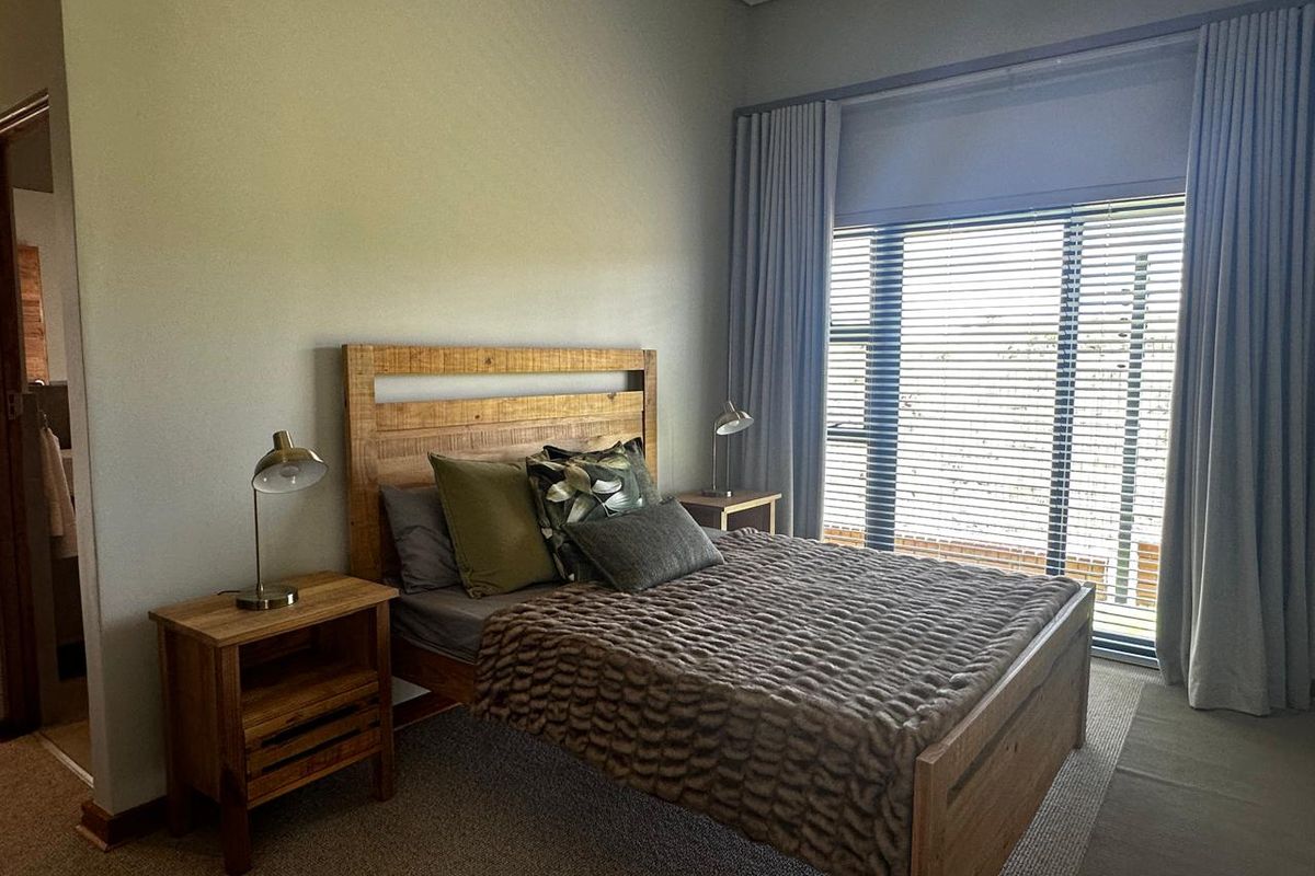 A well-lit bedroom featuring neutral carpeting, large windows with fitted blinds and curtains, and direct access to an adjoining bathroom.
