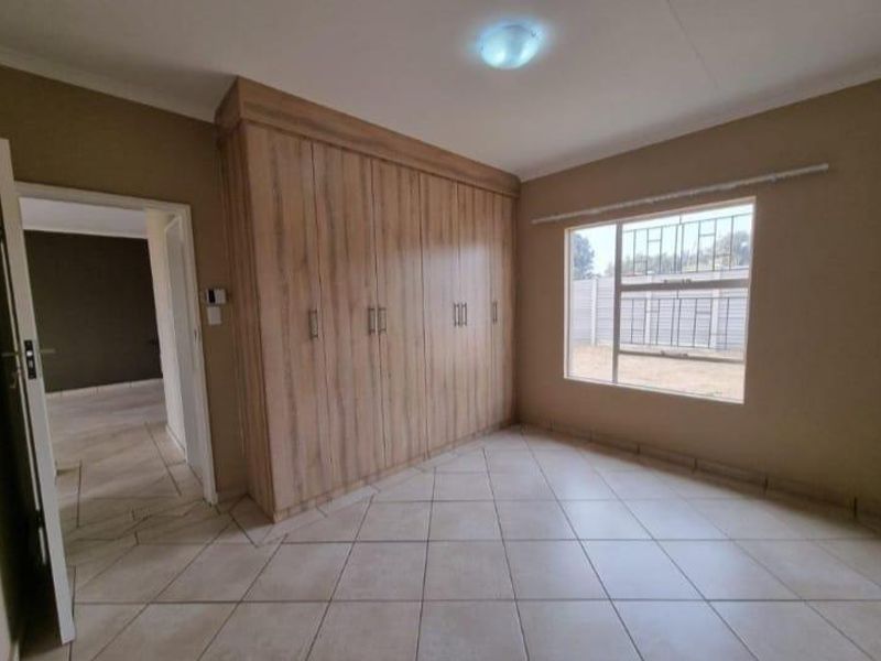 A clean, empty bedroom featuring light wood-finish built-in cupboards, neutral tiled flooring, and a window fitted with security bars.