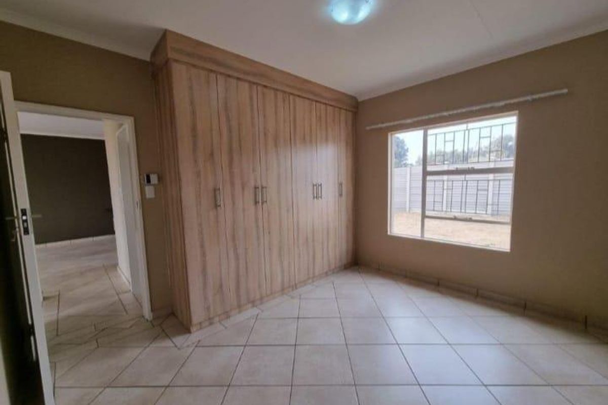 A clean, empty bedroom featuring light wood-finish built-in cupboards, neutral tiled flooring, and a window fitted with security bars.