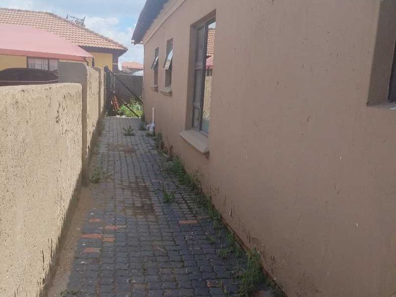A narrow paved side passage along the exterior of the house, featuring a boundary wall and aluminium window frames.