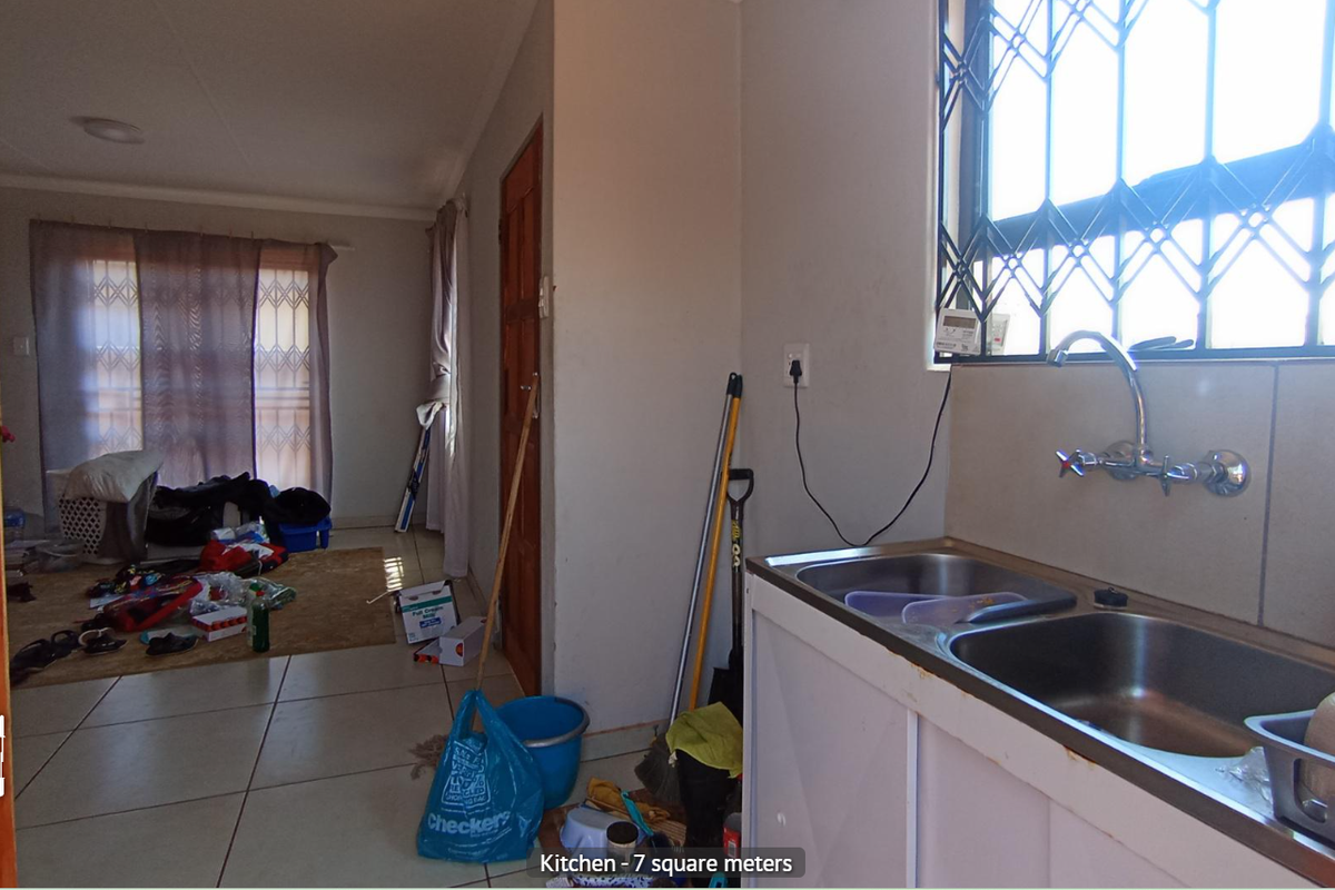 A compact kitchen area featuring a double stainless steel sink, tiled flooring, and security bars on the window, leading into an adjacent living space.