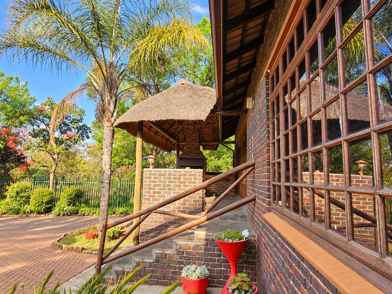 A covered outdoor patio area featuring a traditional thatched roof, built-in brick braai, and classic brickwork, overlooking a landscaped garden.