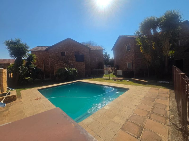 Communal swimming pool area featuring a paved deck, brick apartment building facades, and established garden surroundings.