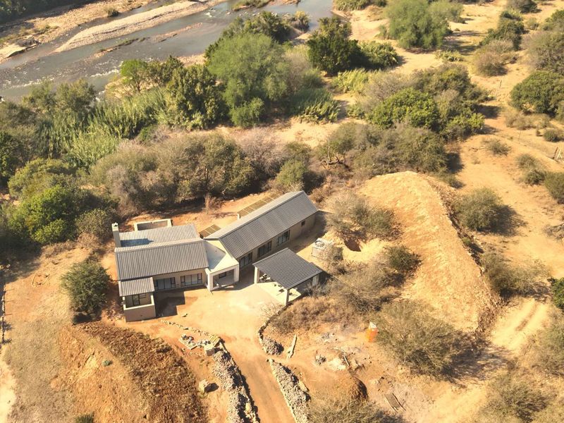 Aerial view of a modern house with a metal roof, situated in a rural landscape near a river, featuring a driveway and covered patio area.