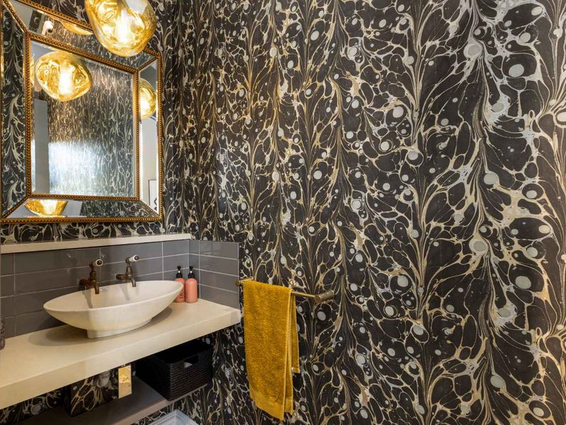 A stylish powder room featuring a vessel sink, stone vanity countertop, grey tiled backsplash, and bold patterned wallpaper.