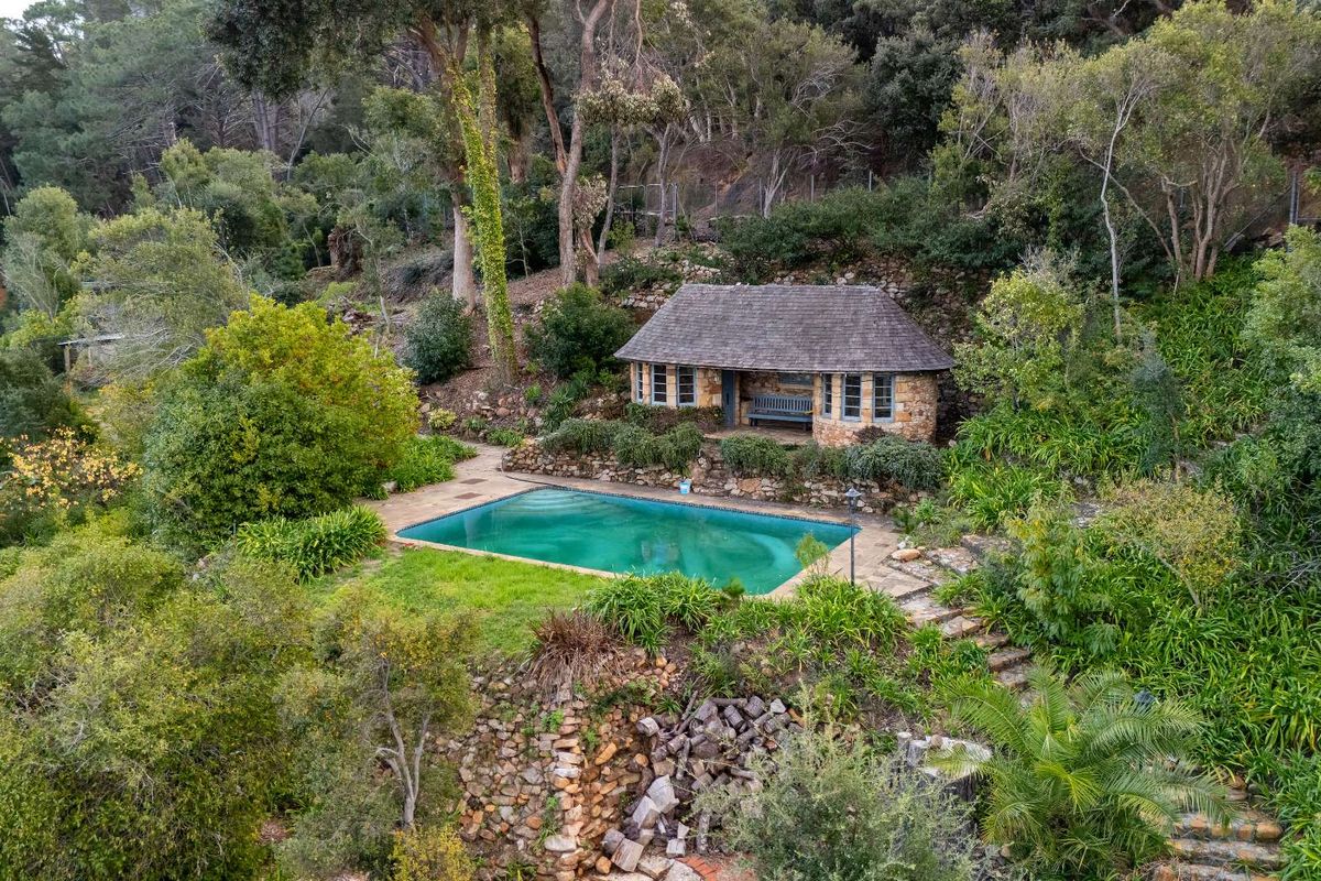 Aerial view of a secluded residential property featuring a swimming pool and a stone-built pool house nestled within a lush, wooded garden setting.