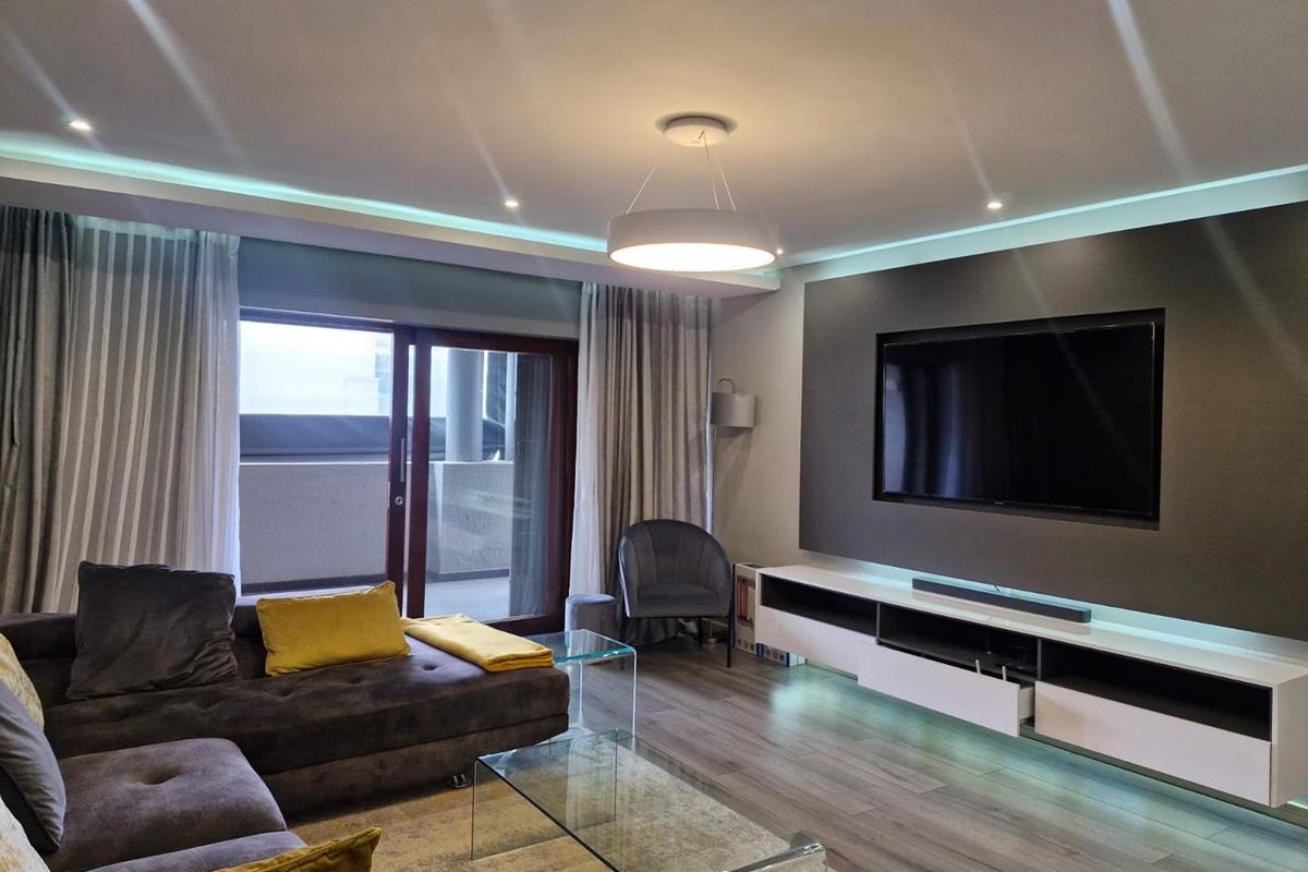 Modern living room featuring recessed ceiling lighting, ambient LED strip lighting, and a built-in media console with sliding glass door access to the balcony.