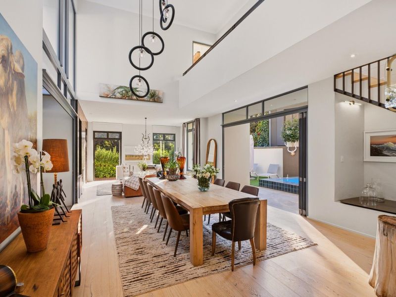 Spacious open-plan dining area featuring double-volume ceilings, modern pendant lighting, light timber flooring, and seamless flow to an outdoor patio and pool area.