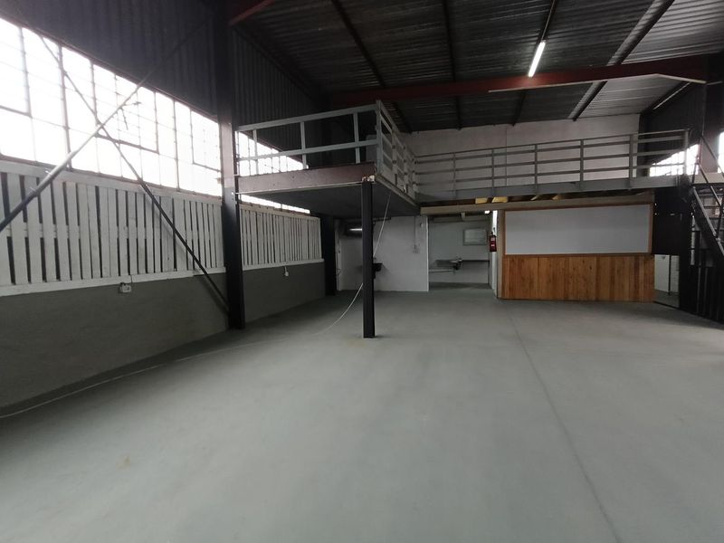Spacious industrial open-plan warehouse area featuring a mezzanine level, high ceilings with exposed beams, large windows, and durable concrete flooring.