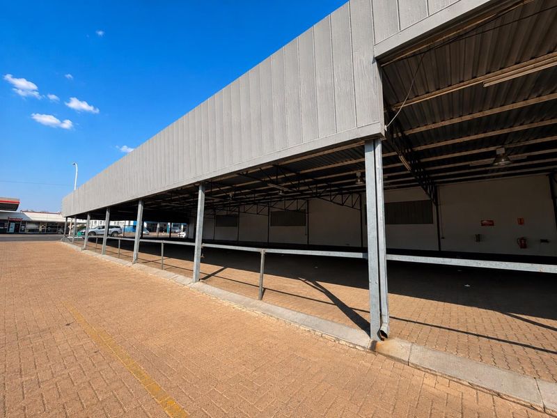 A wide exterior view of a commercial property featuring a large covered parking or loading area with a paved surface and steel structural supports.
