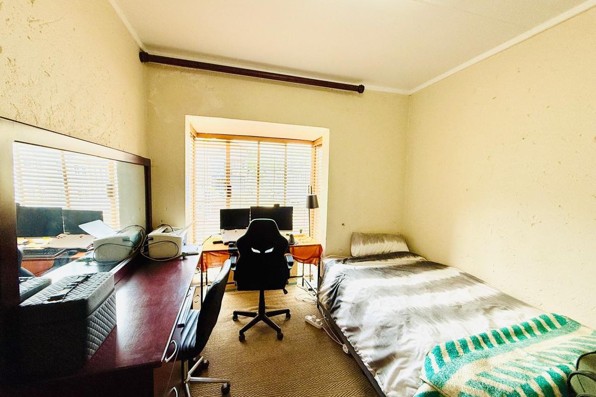A compact bedroom featuring a window with fitted blinds and carpeted flooring, currently utilized as a combined sleeping and workspace area.