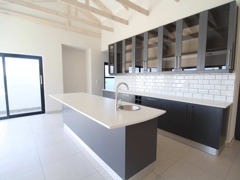 Modern kitchen featuring a central island, sleek dark cabinetry with glass-front uppers, subway tile backsplash, and exposed timber trusses under a vaulted ceiling.