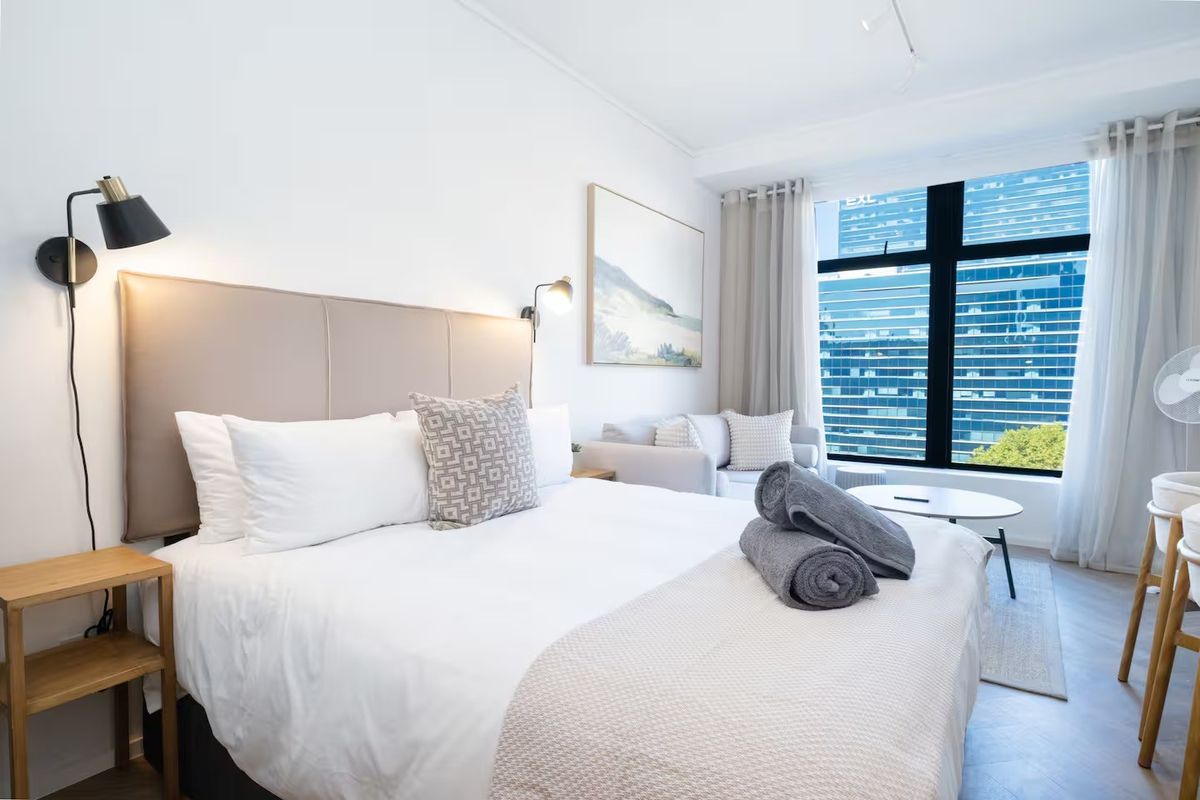 A bright and modern bedroom featuring large windows with city views, herringbone flooring, and contemporary wall-mounted lighting fixtures.