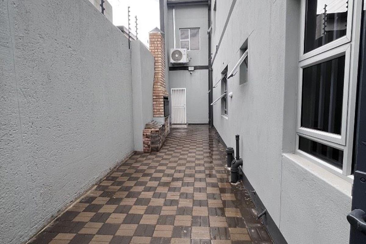 A narrow paved side passage featuring a built-in brick braai, secure electric fencing, and external wall-mounted air conditioning unit.