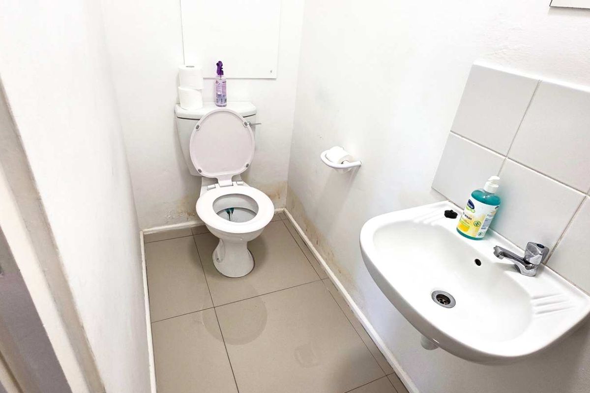 A compact office restroom featuring a wall-mounted basin, tiled splashback, and standard toilet, currently showing signs of wear and water staining on the walls.