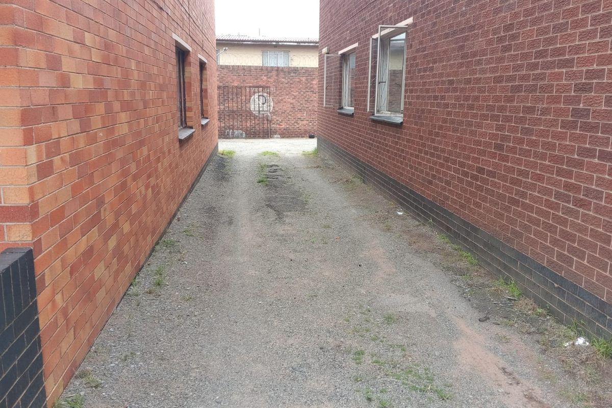 A narrow gravel driveway situated between two brick apartment building facades, leading to a rear gated entrance.