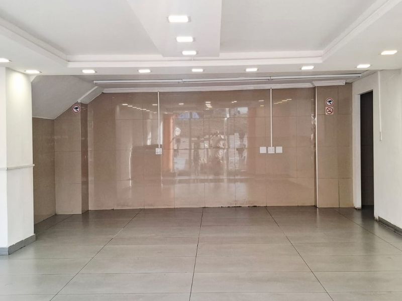 A spacious, empty open-plan commercial area featuring neutral tiled flooring, a tiled feature wall, and modern recessed ceiling lighting.
