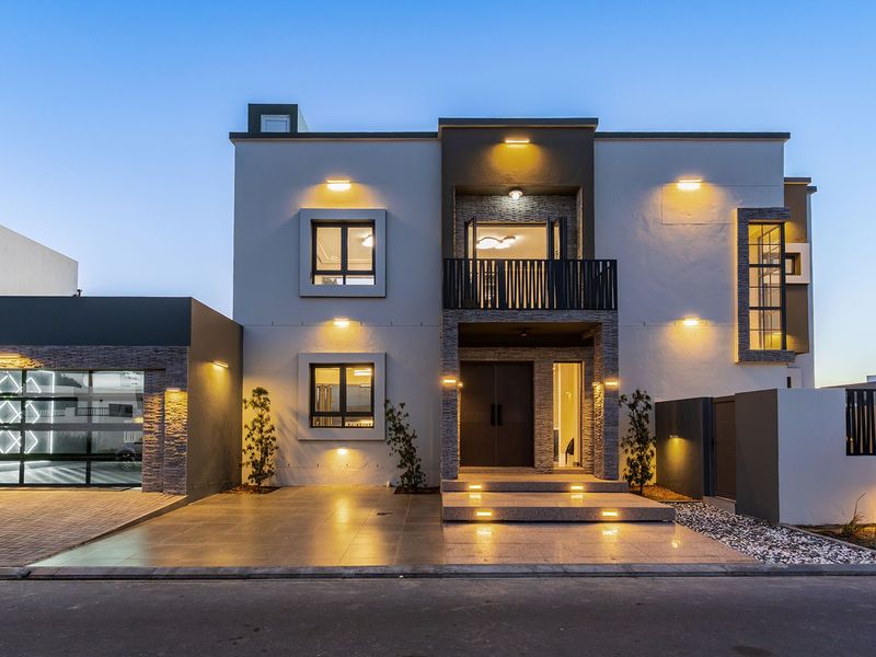 Modern double-story residential facade featuring stone cladding, a balcony, integrated lighting, and a spacious paved driveway with a double garage.
