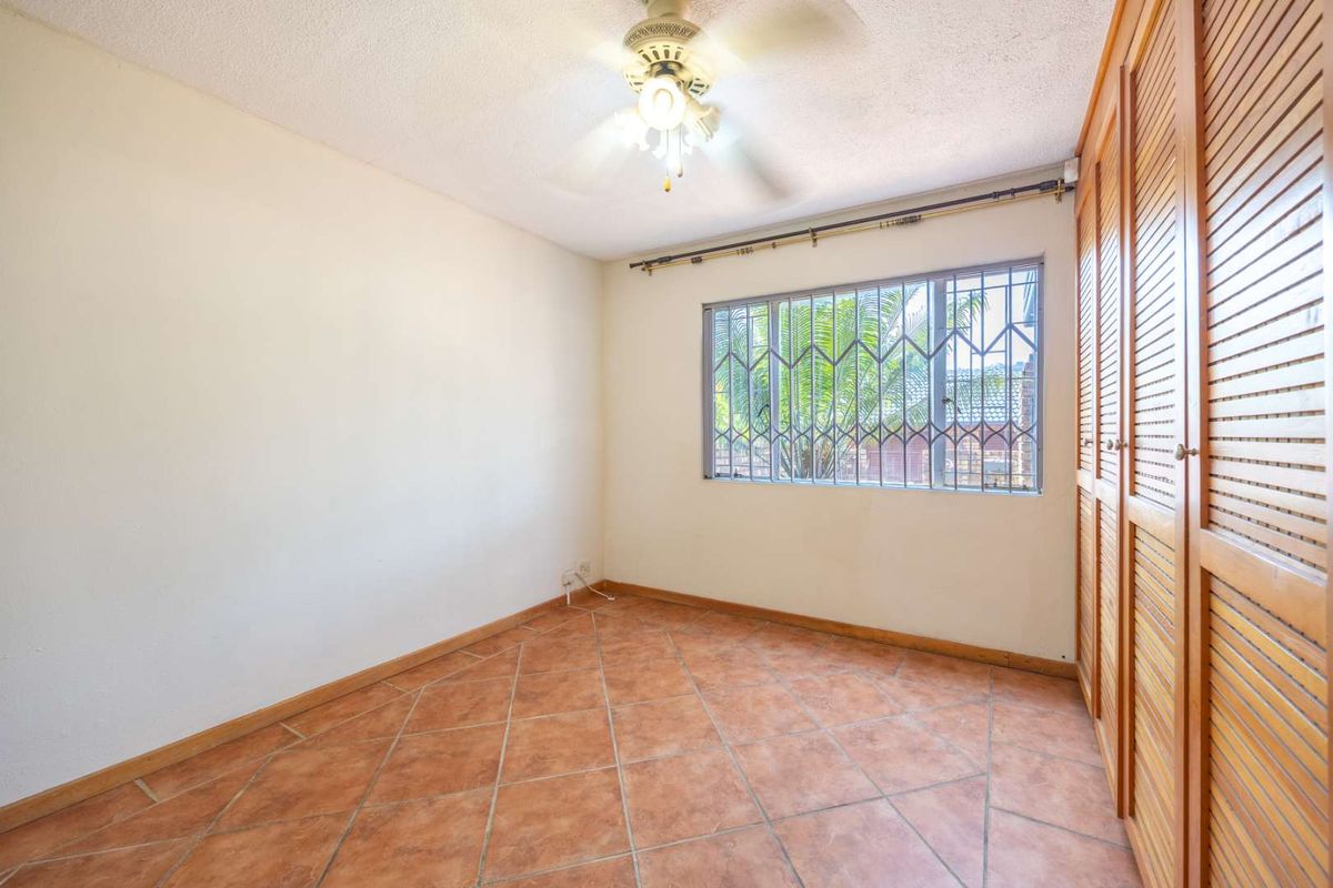 A vacant bedroom featuring tiled flooring, built-in wooden cupboards, a ceiling fan, and a window fitted with security bars.