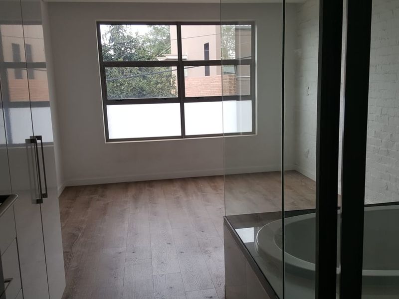 A modern, empty bedroom featuring laminate flooring, built-in cupboards, and an integrated en-suite bathtub behind a glass partition.