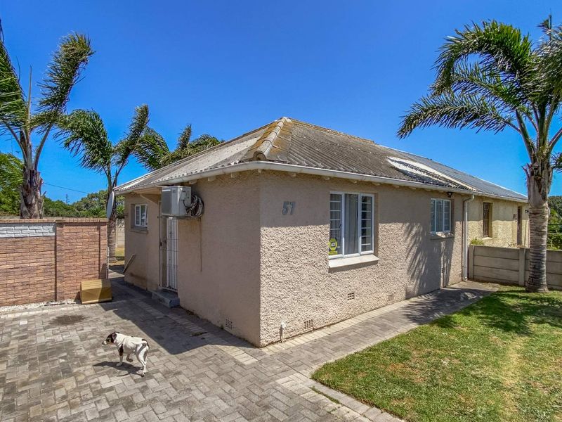 Rear exterior view of a single-story house featuring a paved driveway, boundary wall, and established palm trees under a clear blue sky.