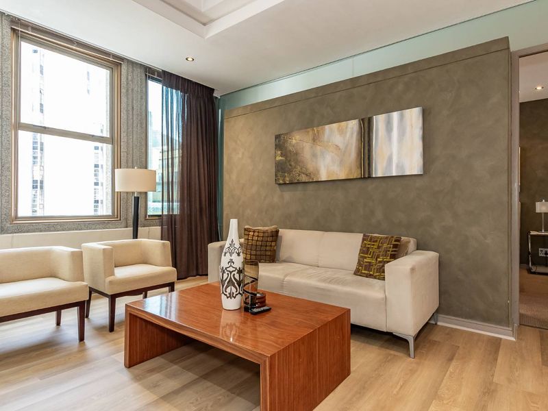 A well-lit living room featuring modern laminate flooring, a textured feature wall, and large windows providing ample natural light.