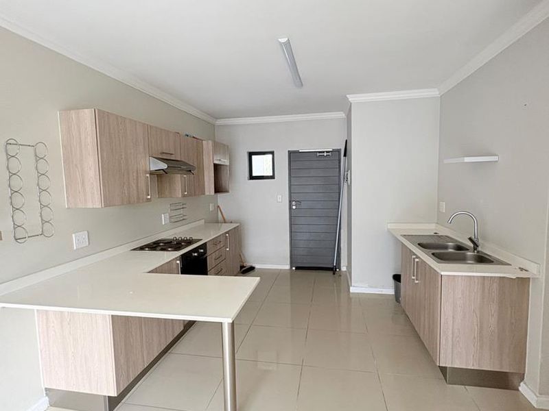 A modern, open-plan kitchen featuring light wood-finish cabinetry, stone countertops, a breakfast bar, and integrated appliances, set against neutral tiled flooring.