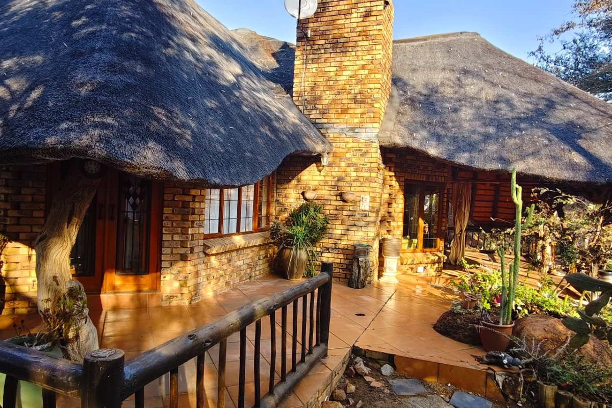 A rustic exterior view featuring a traditional thatched roof, brick chimney, and a tiled patio area surrounded by natural landscaping.