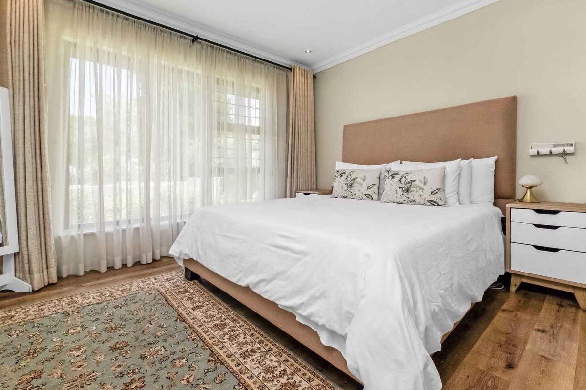 A well-maintained bedroom featuring large windows with sheer curtains, crown molding, and warm-toned wood flooring.
