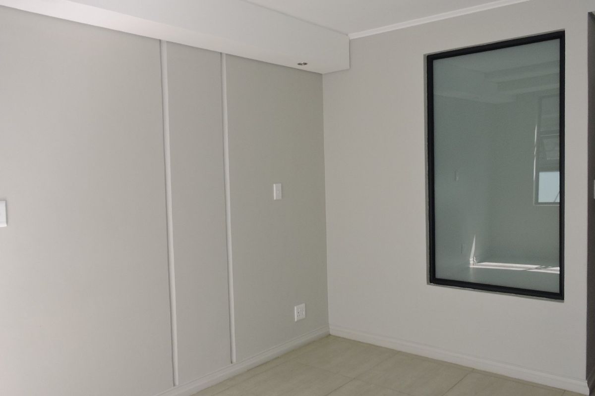 A clean, neutral-toned bedroom featuring tiled flooring, a built-in wardrobe, and a modern black-framed window, offering a bright and versatile living space.