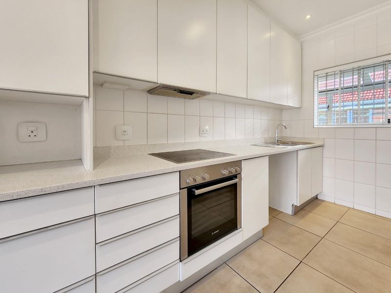 A clean, modern kitchen featuring white cabinetry, stone countertops, integrated oven and hob, and tiled finishes, offering a functional and bright workspace.