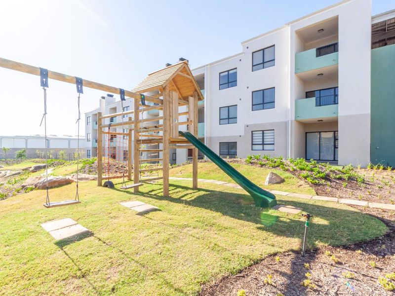 A well-maintained communal garden area featuring a wooden playground set, situated in front of a modern apartment complex with balconies.